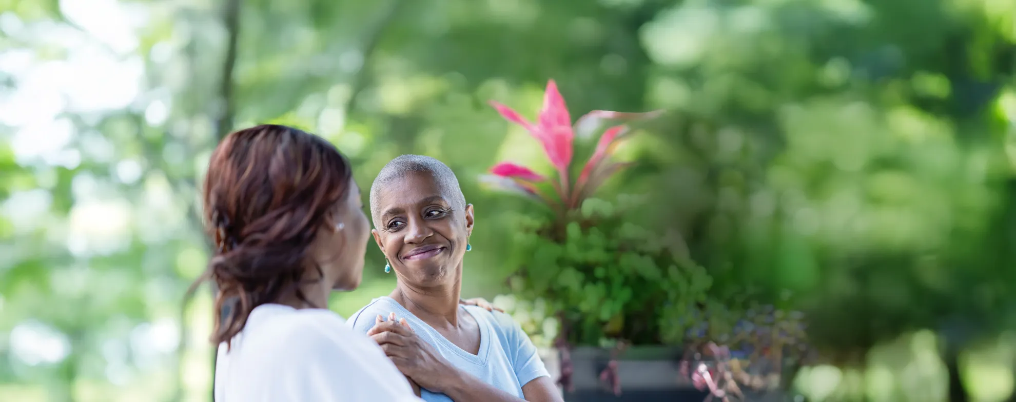 Two women outdoors, one with a shaved head, smiling and holding hands in a supportive moment.